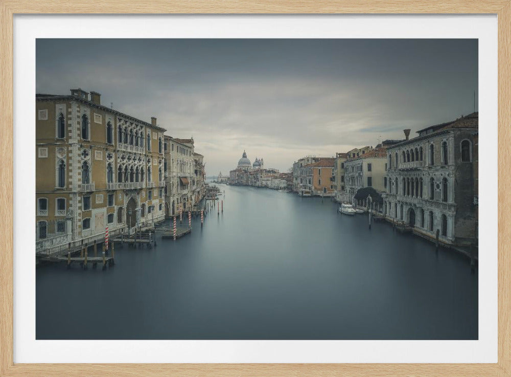 A long-exposure photograph of the Grand Canal in Venice, Italy, featuring silky smooth dark water flowing between historic, ornate buildings under a cloudy, overcast sky. The image is presented in a silver frame. Artwork