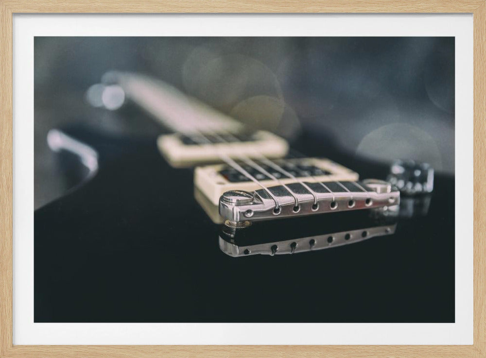 A close-up, framed photograph of a black electric guitar with a shallow depth of field. The focus is sharp on the chrome bridge and its reflection on the glossy black body. The pickups and fretboard are blurred in the background with soft bokeh lights. Decor