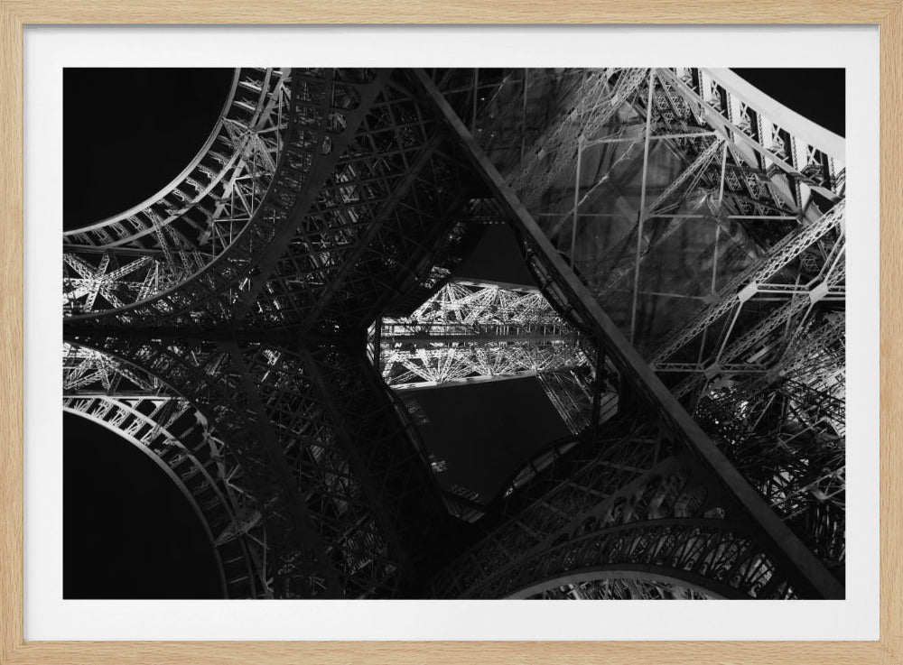 A dramatic black and white photograph shot from a low angle, looking up through the intricate iron latticework of the Eiffel Tower at night. The illuminated structure creates a complex, almost abstract pattern of beams, arches, and trusses against the dark night sky. The image is presented within a silver-colored frame. Wall Art