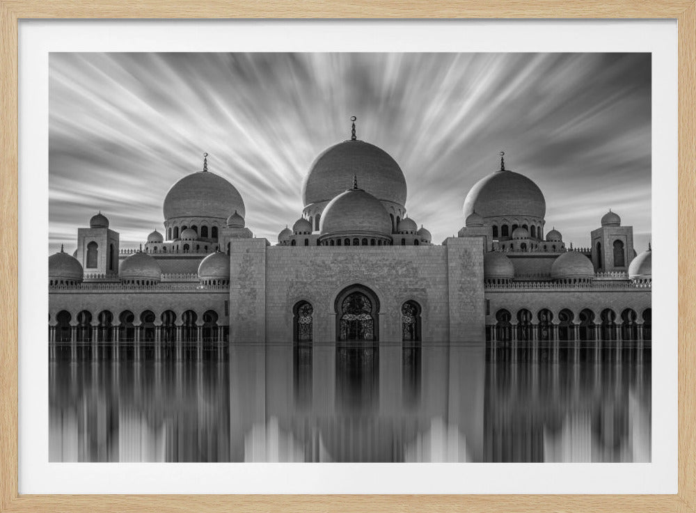 A stunning black and white long-exposure photograph of a grand mosque with multiple domes, reflected perfectly in calm water. The sky above is dramatic, with clouds streaking outwards, and the image is presented in a silver frame. Decor