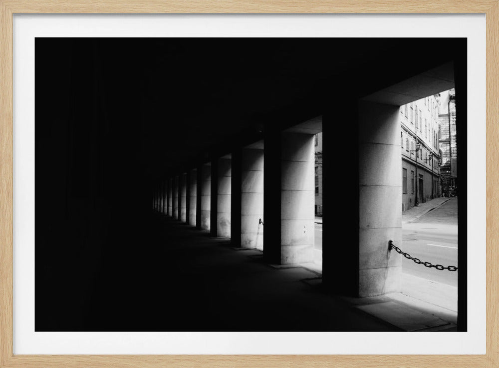 A high-contrast, black and white photograph from the perspective of a dark, covered walkway lined with a row of thick pillars. Light from an adjacent street highlights the pillars, creating a dramatic play of light and shadow and a strong sense of depth. Print
