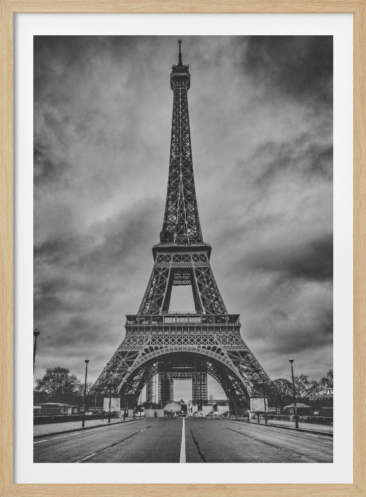 A dramatic, low-angle black and white photograph of the Eiffel Tower in Paris. The view is from the center of a road leading towards the iconic landmark, set against a backdrop of a cloudy, moody sky and framed in black. Artwork