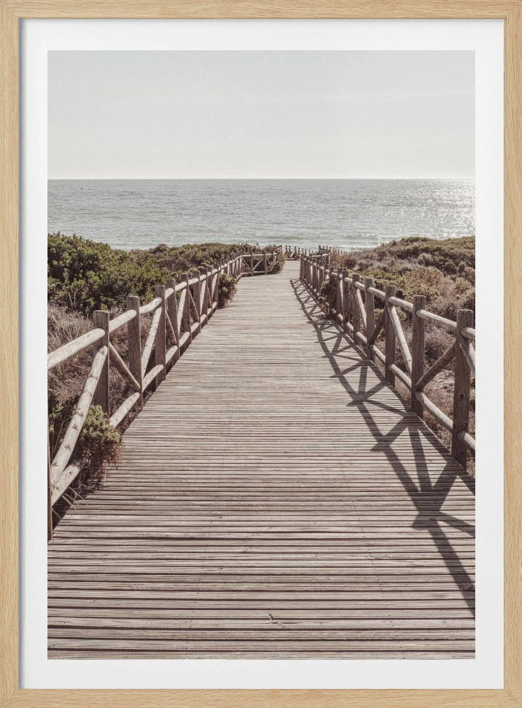 A long wooden boardwalk with rustic log railings leads the viewer's eye towards a calm, sunlit ocean under a hazy sky. Strong shadows from the railings fall across the path, creating a striped pattern. Poster