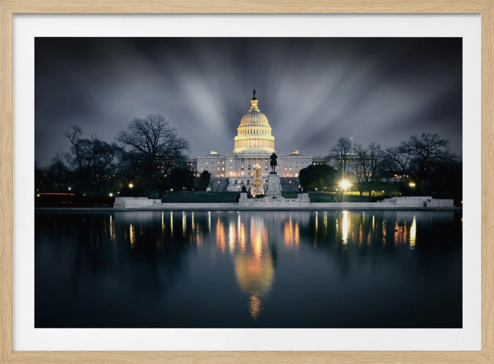 A dramatic long-exposure night photograph of the illuminated US Capitol Building, with its dome glowing brightly against a dark grey sky. The building and its lights are perfectly reflected in the still water of the reflecting pool in the foreground. The image is presented in a silver frame. Decor