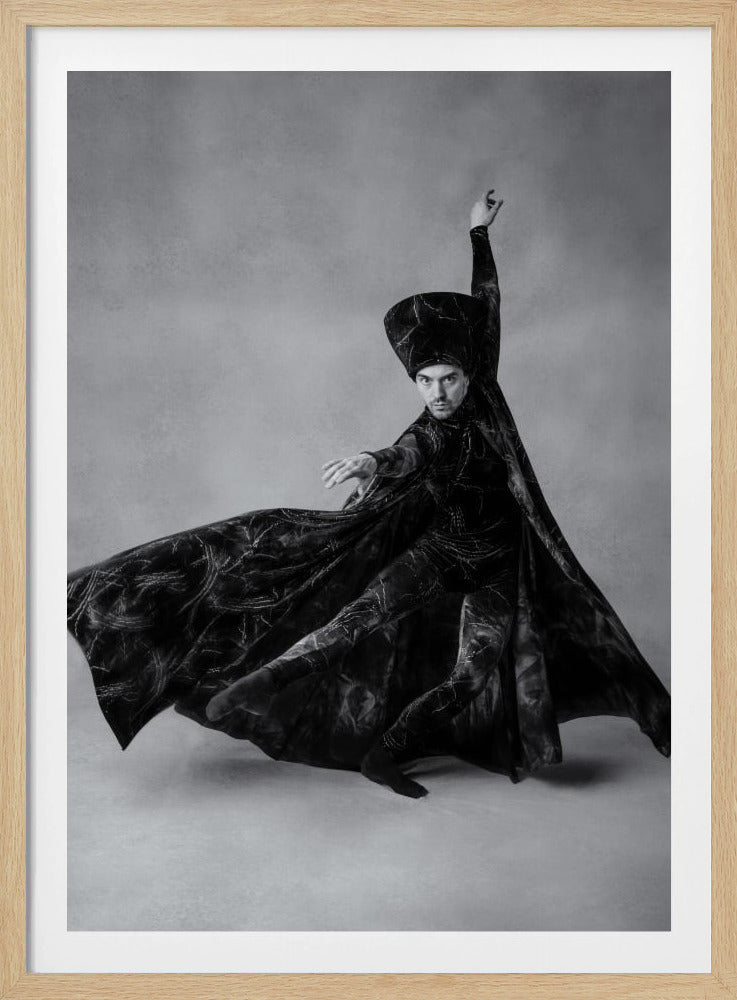 A dramatic black and white photograph of a male dancer in mid-performance. He wears an elaborate, dark, patterned costume with a flowing cape and a tall, structured hat. His body is captured in a dynamic pose, with arms extended and the cape swirling around him, emphasizing the sense of movement. He looks directly at the viewer with an intense and focused expression against a plain gray studio background. Wall Art