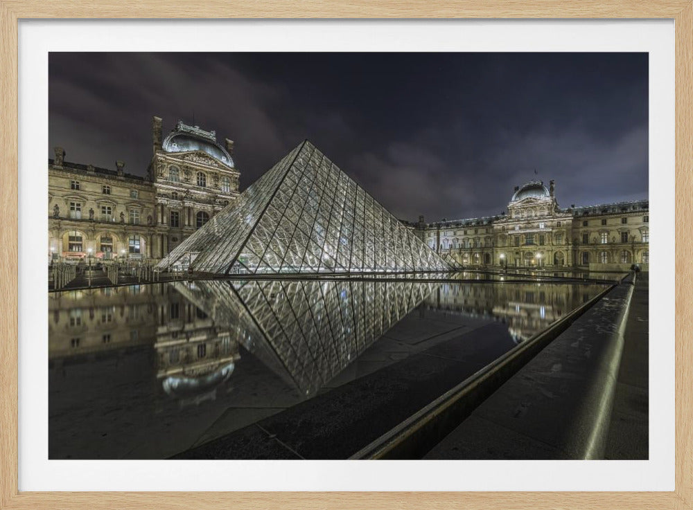 A dramatic night photograph of the illuminated Louvre Pyramid in Paris, its glass structure glowing against a dark, cloudy sky. The historic Louvre Palace is visible in the background, and the entire scene is perfectly reflected in the still water of a foreground pool. The image is presented within a silver frame. Artwork