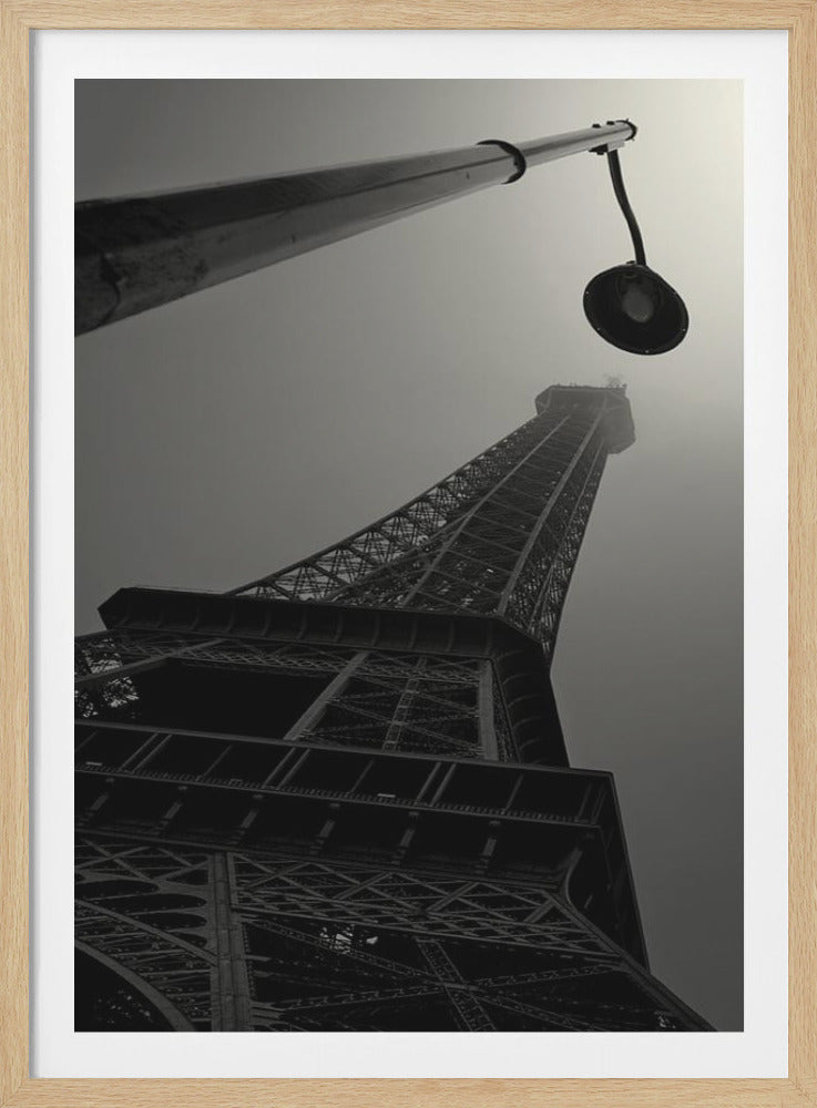 A dramatic low-angle, black and white photograph of the Eiffel Tower in Paris, with a street lamp in the foreground pointing towards the iconic structure as it disappears into the misty sky. Artwork