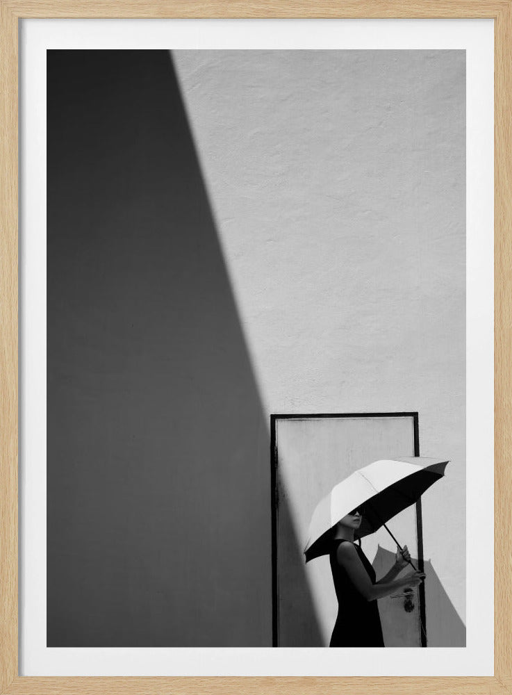 A minimalist black and white photograph of a woman in a black dress holding a two-toned umbrella. She stands in front of a door against a light wall, with a sharp, diagonal shadow dramatically cutting across the upper left of the image, creating a high-contrast, geometric composition. Decor