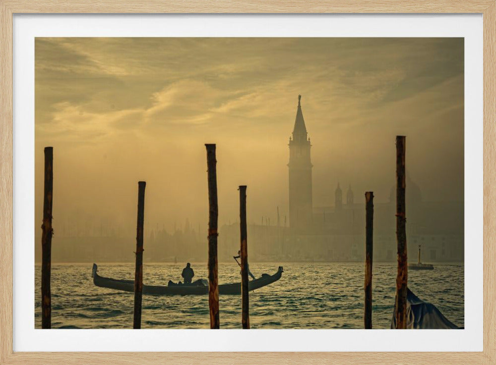 A lone gondolier navigates a canal in Venice during a hazy, golden sunrise. In the foreground, wooden poles rise from the water, and a historic bell tower is silhouetted in the misty background. The image is presented within a silver frame. Print