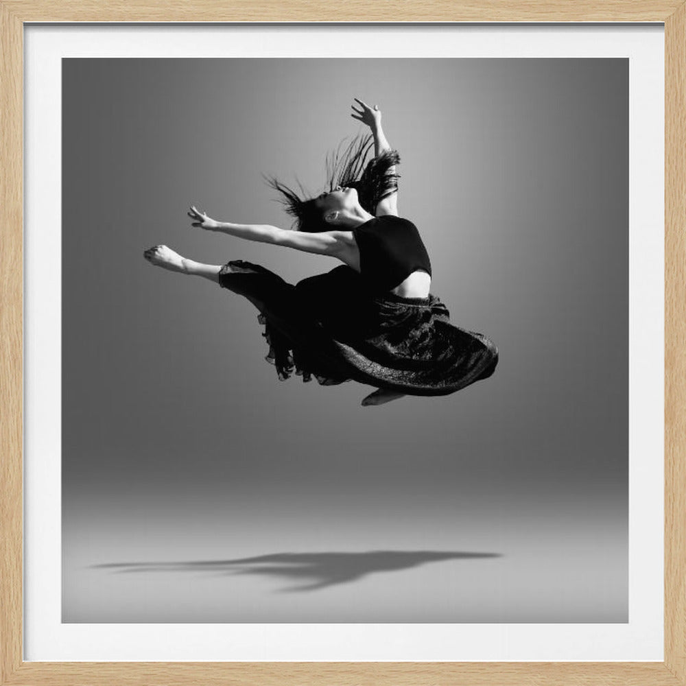 A framed, black and white photograph of a graceful female dancer captured mid-leap against a neutral gray background. She wears a black crop top and a flowing skirt, with her back arched, arms extended, and long dark hair flying through the air, conveying a sense of freedom and movement. A soft shadow is cast on the floor below her. Artwork