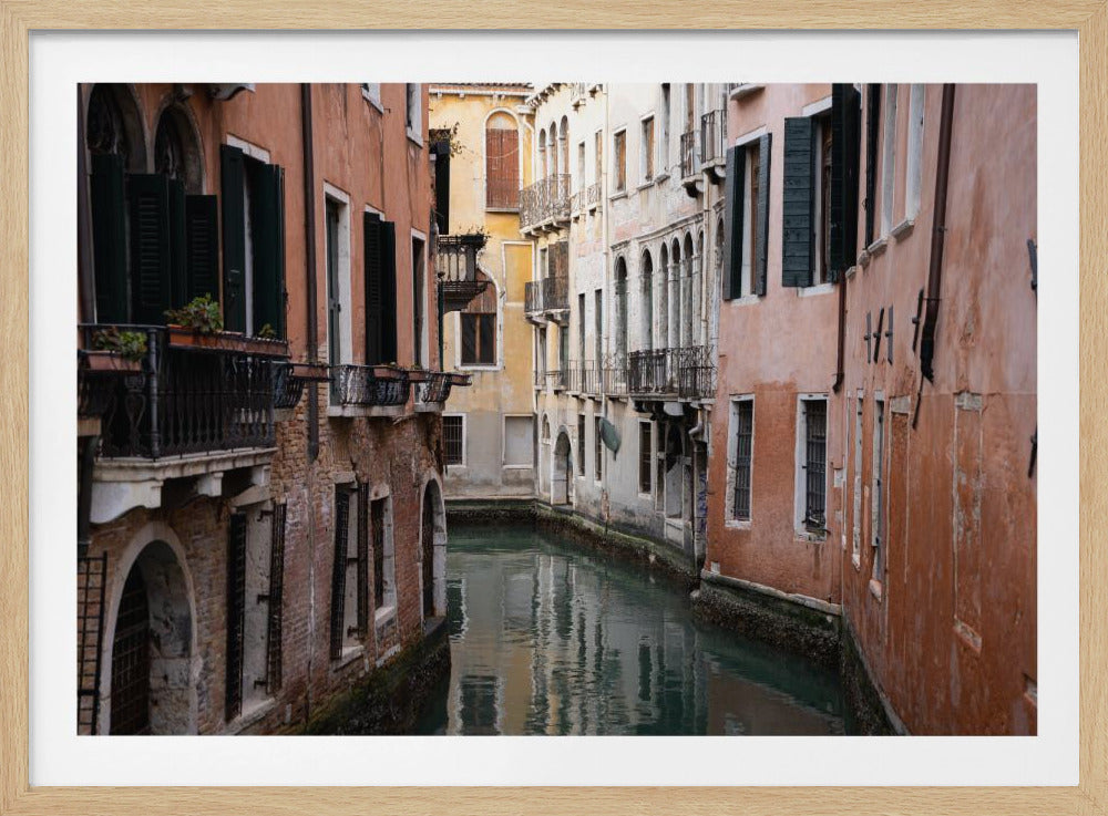 A framed photograph of a narrow canal in Venice, Italy, enclosed by tall, historic buildings with weathered terracotta and peach-colored facades, dark shutters, and wrought-iron balconies. The calm, dark water of the canal reflects the surrounding architecture. Artwork