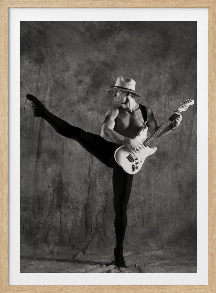 A dramatic black and white studio portrait of a shirtless, muscular man wearing a fedora and black tights. He holds a white electric guitar while gracefully balancing on one leg, with his other leg extended high in a ballet pose. Wall Art