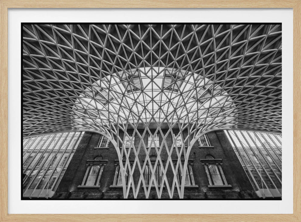 A framed, black and white photograph of the Western Concourse at King's Cross Station, London. The view is from a low angle, looking up at the intricate, white, funnel-like lattice roof structure as it spreads out across the ceiling, contrasting with the classic brick facade of the building behind it. Decor