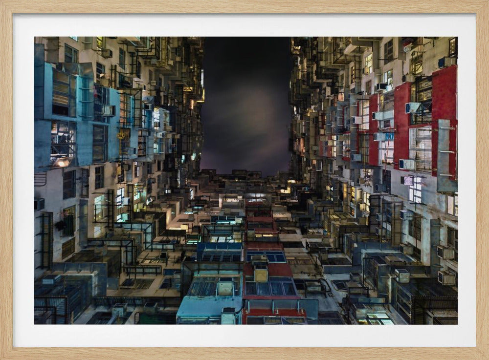 A framed photograph from a low-angle perspective, looking up between two towering, densely packed apartment buildings at night. The buildings are a collage of colors and textures with many windows lit up, creating a claustrophobic yet captivating urban scene against a dark sky. Wall Art
