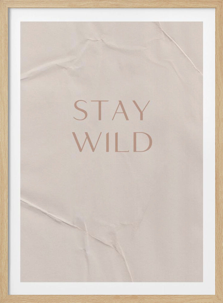 A minimalist poster in a thin black frame. The words 'STAY WILD' are written in a dusty rose, elegant font at the center. The background is a textured, light beige paper that appears creased and folded. Print