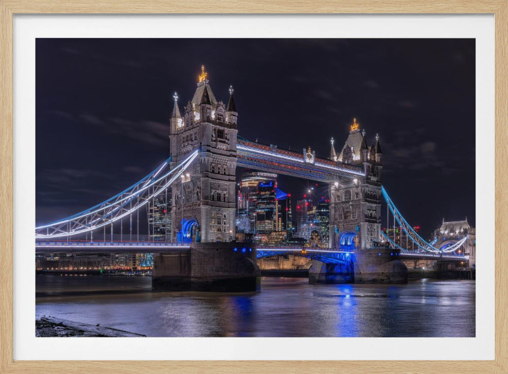 A vibrant night photograph of the iconic Tower Bridge in London, illuminated with bright white and electric blue lights. The dark River Thames in the foreground reflects the bridge and the lit-up modern city skyline in the background, all under a dark, cloudy sky. The image is enclosed in a silver frame. Decor