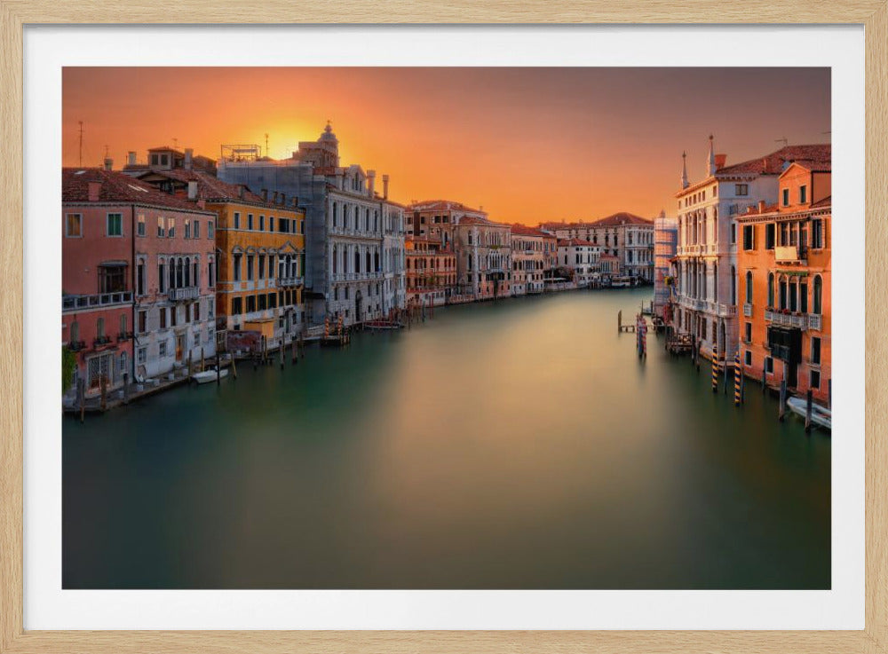 A long-exposure photograph of the Grand Canal in Venice at sunset, captured in a silver frame. The sky is a brilliant orange, reflecting on the smooth, glassy surface of the canal. Ornate, historic buildings line both sides of the water, bathed in the warm light of the setting sun. Poster