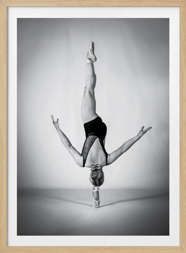 A black and white photograph of a ballerina performing an incredible feat of balance and strength. She is upside down, doing a handstand on the point of one ballet shoe, with her head also on the floor. Her other leg is extended straight up into the air, and her arms are spread out to the sides for balance. The photo is framed with a black border. Poster
