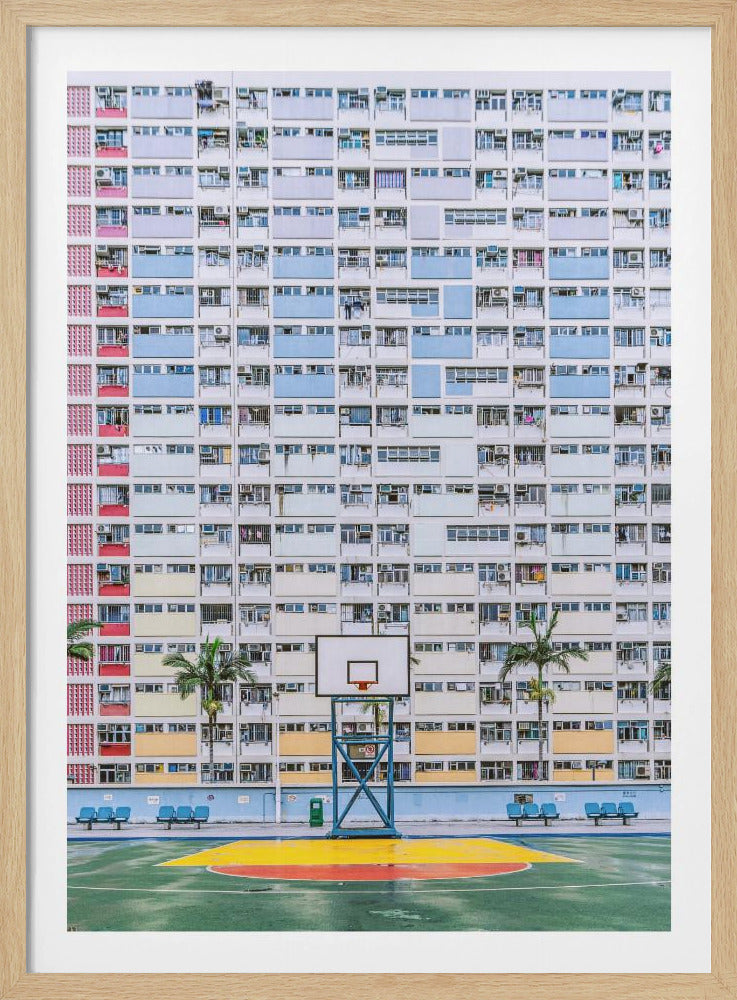 A symmetrical photograph of a massive, multi-colored apartment building with a basketball court in the foreground. The building's facade is a dense grid of windows and balconies, painted in white and pastel shades of blue, red, and yellow. Below, a single basketball hoop stands on the green court, which has a yellow and red painted key. Decor