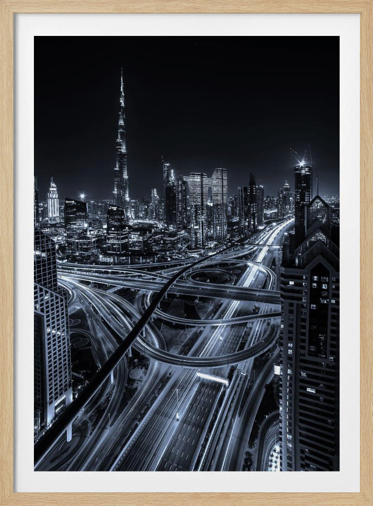A cool-toned black and white photograph of the Dubai skyline at night. The iconic Burj Khalifa stands tall in the background, while the foreground is dominated by a massive, complex highway interchange with light trails from moving cars, creating a sense of motion and modernity. Poster