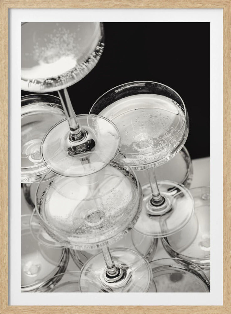 A close-up, high-angle photograph in black and white of a champagne tower. Several coupe-style glasses are stacked, filled with bubbly champagne that catches the light. The background is dark, creating a strong contrast and highlighting the elegant glassware. Decor