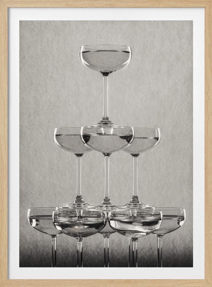A classic black and white photograph of a champagne tower, with coupe glasses stacked in a pyramid. The glasses are filled with bubbly liquid, and the composition is set against a textured light gray background and enclosed in a black frame. Poster