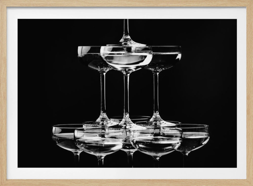 A black and white photograph of a champagne tower made of coupe glasses stacked in a pyramid shape against a solid black background, all within a silver frame. Print