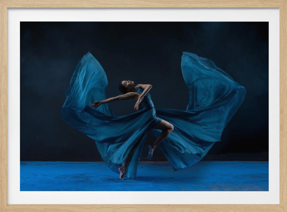 A graceful dancer wearing a long, flowing blue dress is captured mid-pose against a dark background. The dress's fabric billows out on either side of her like giant wings. She is barefoot on a textured blue floor, with one leg bent and arms outstretched in an elegant, expressive movement. The image is presented within a silver frame. Print