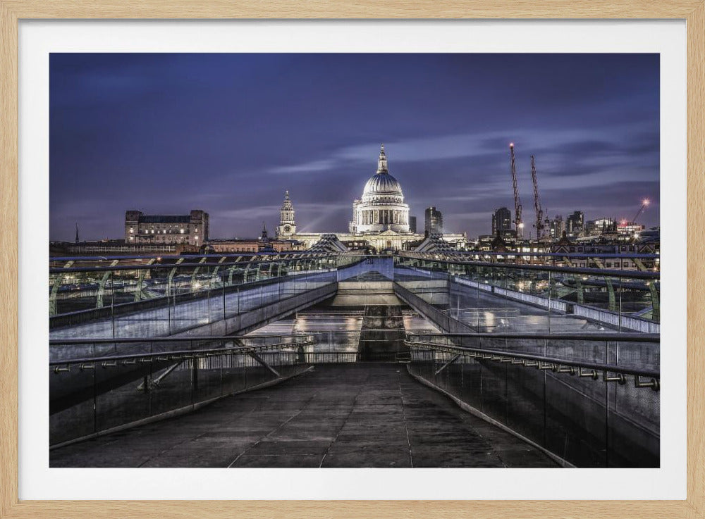 A nighttime photograph from the Millennium Bridge leading towards the brightly illuminated St. Paul's Cathedral against a dark blue twilight sky in London, all enclosed in a silver frame. Poster