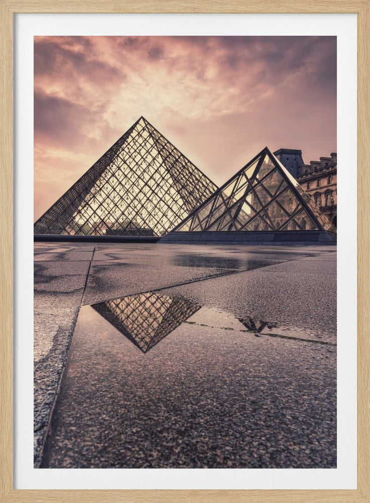 A low-angle photograph of the Louvre Pyramid in Paris, with its glass and metal structure reflected in a puddle on the wet stone ground in the foreground. The sky above is filled with soft, pink and purple clouds at dusk or dawn. Decor