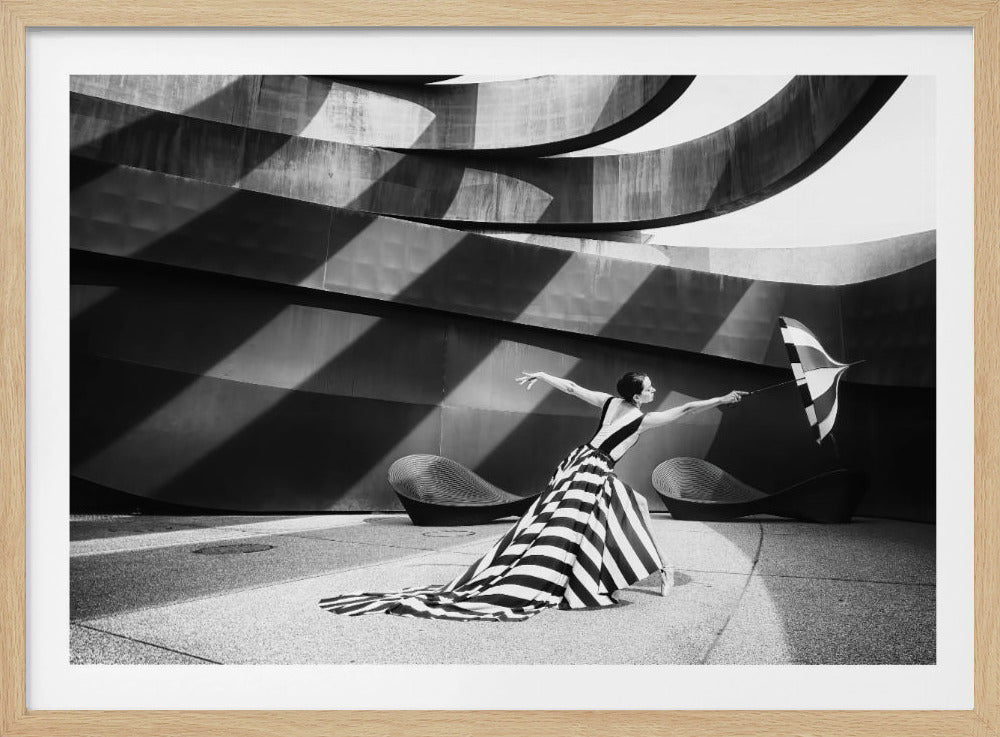 A dramatic black and white photograph of a woman in a long, flowing black-and-white striped gown. She strikes a dynamic, dance-like pose, holding a matching striped umbrella against an angular, modern architectural background with strong diagonal shadows. Decor