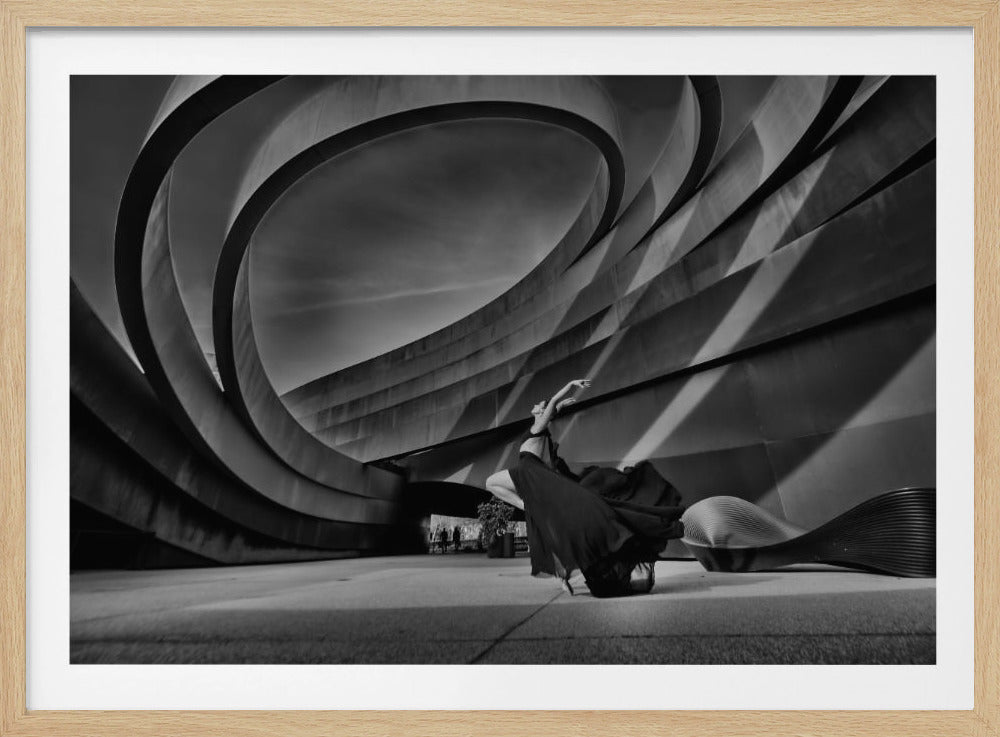 A dramatic black and white photograph from a low angle, showing a graceful dancer in a flowing black dress posing in front of a monumental, modern building with sweeping, curved walls and striking shadows. The image is presented within a silver picture frame. Wall Art