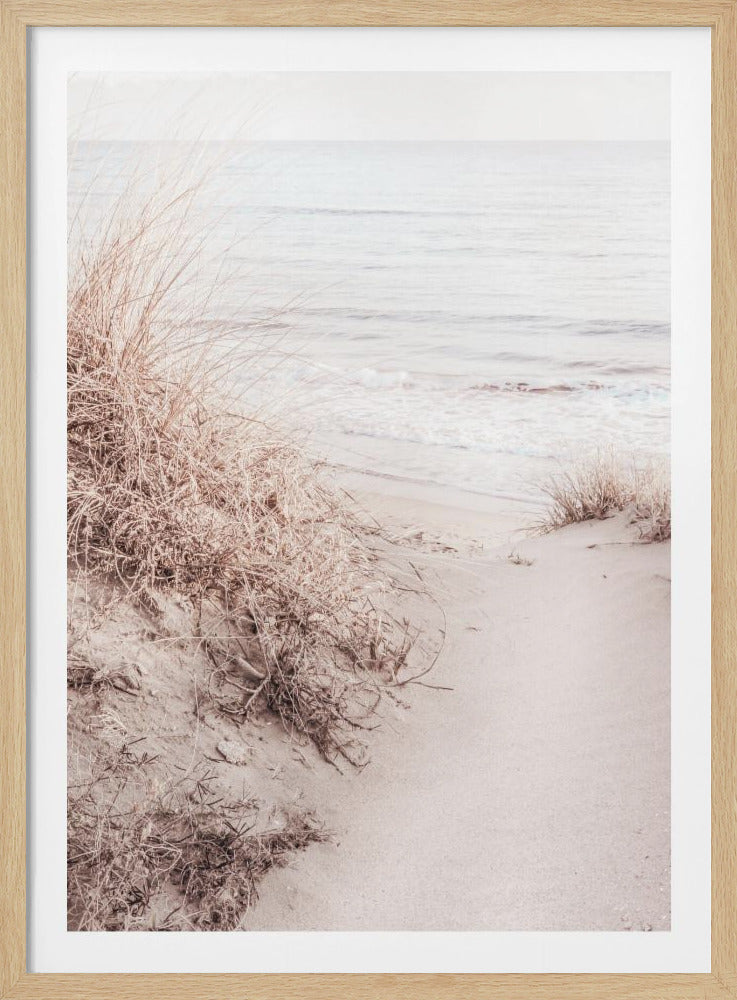 A tranquil beach scene with pale, dried grasses on a sand dune in the foreground, leading down to the calm, light-colored ocean under a hazy sky. Wall Art