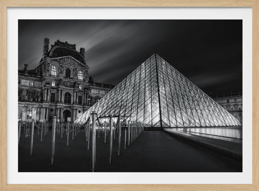 A dramatic black and white photograph of the Louvre Pyramid in Paris at night. The glass pyramid is brightly illuminated from within, creating a stark contrast with the dark, streaky sky. Part of the historic Louvre Palace is visible in the background, and metal stanchions with ropes line the foreground. Artwork