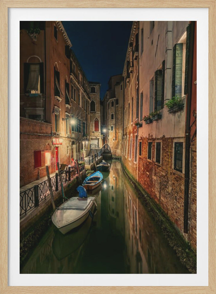 A tranquil night scene of a narrow canal in Venice, Italy. Old, warmly lit buildings line the dark water, with several small boats moored along the sides, their reflections shimmering on the surface under a dark night sky. Decor