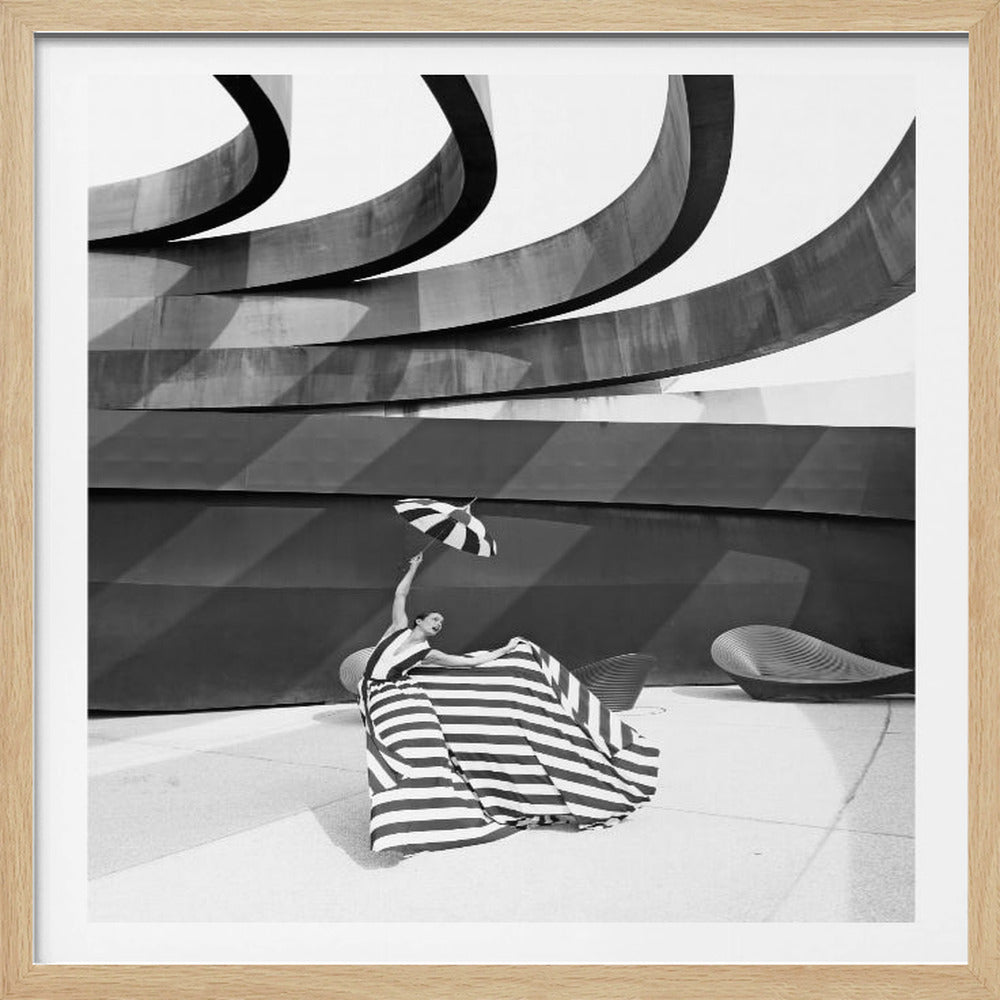 A framed, black and white photograph of a woman in a dramatic, horizontally striped gown, holding a matching striped parasol aloft against a modern, abstract architectural background with bold curves and shadows. Print