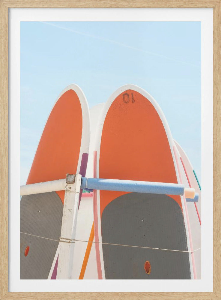 A close-up shot of several orange and white paddleboards standing vertically in a rack against a clear, light blue sky on a sunny day. Decor