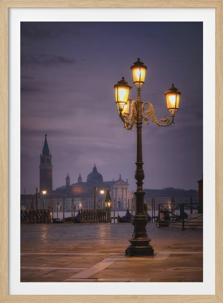 An ornate, three-headed street lamp glows warmly in the foreground against a dusky purple sky in Venice. In the background, across the water, the iconic silhouette of the Church of San Giorgio Maggiore and its campanile can be seen, with gondolas moored along the wet, reflective plaza. Wall Art
