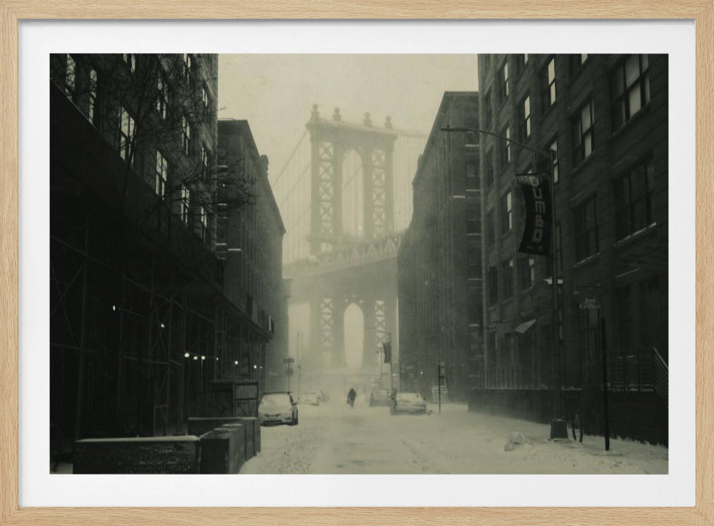 A framed, vintage-style black and white photograph capturing a snowy day in DUMBO, Brooklyn. The view looks down a snow-covered street lined with dark buildings, perfectly framing the Manhattan Bridge in the foggy distance. A few cars are parked along the street, and snow is visibly falling, creating a quiet, atmospheric winter scene. Decor