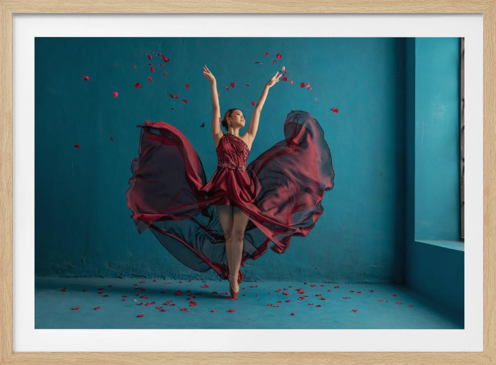 A graceful ballerina in a flowing dark red dress dances on pointe against a solid teal-blue background. Her arms are raised elegantly as red rose petals rain down around her and scatter on the floor. The entire image is presented within a textured silver frame. Poster