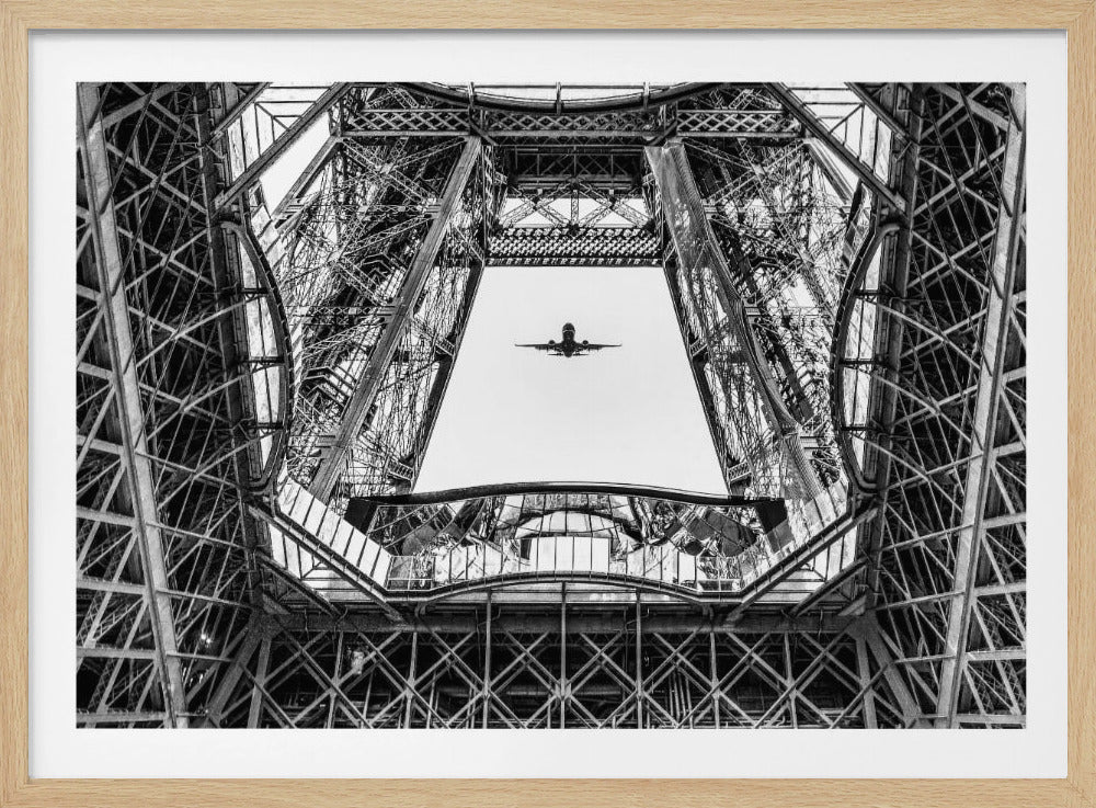 A framed, black and white, low-angle photograph looking up through the intricate steel lattice of the Eiffel Tower, perfectly framing an airplane flying overhead against a bright sky. Decor