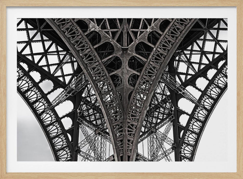 A framed, black and white, low-angle photograph capturing the intricate and symmetrical iron lattice work of the Eiffel Tower. The detailed girders, rivets, and decorative arches create a complex geometric pattern against a bright white sky. Wall Art