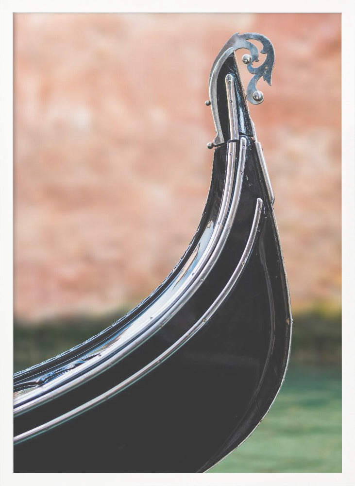 A close-up, vertical photograph of the front prow of a classic black Venetian gondola. The focus is on the shiny, silver, ornate 'ferro' ornament at the tip. The background is a soft-focus blend of a terracotta-colored wall and the green water of a canal. Artwork