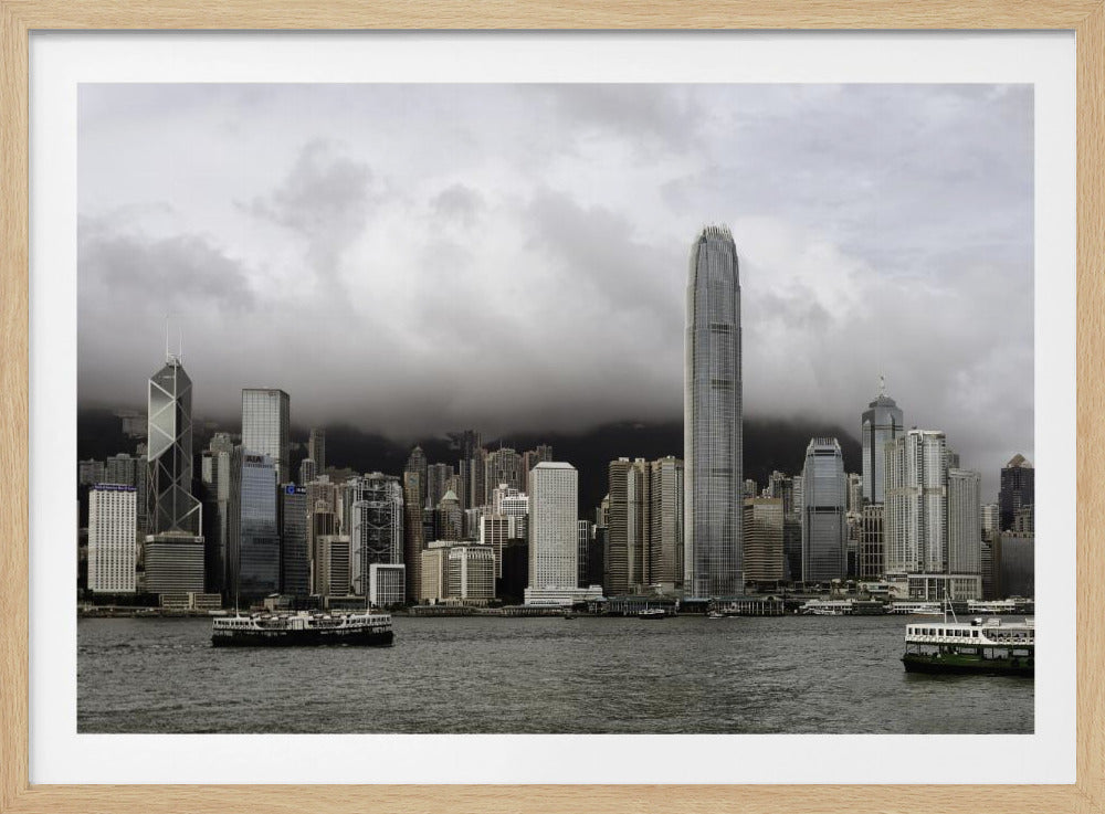 A framed photograph of the Hong Kong skyline on a very cloudy, overcast day. A very tall, modern skyscraper stands out in the center, with the dark water of Victoria Harbour and a green and white ferry in the foreground. Wall Art