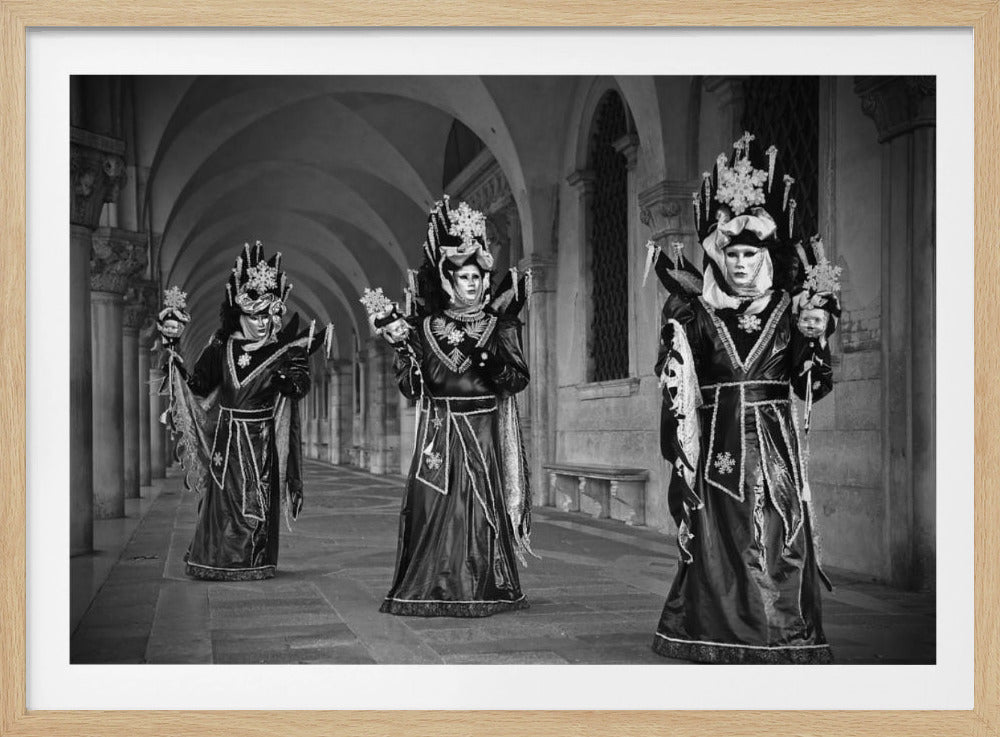 A black and white photograph of three people in elaborate, identical costumes and masks standing in a stone arcade. Their dark outfits are detailed with white patterns, and their headdresses feature large snowflake designs and dangling icicles. The figures hold small masked scepters, creating a mysterious and theatrical scene reminiscent of the Venice Carnival. Wall Art