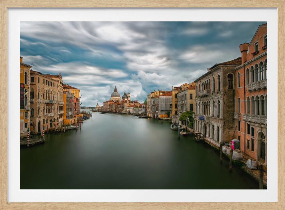 A long-exposure photograph of the Grand Canal in Venice, with smooth, dark water flowing between historic, colorful buildings. The Basilica di Santa Maria della Salute is visible in the distance under a dramatic, swirly sky. The photo is enclosed in a silver frame. Decor