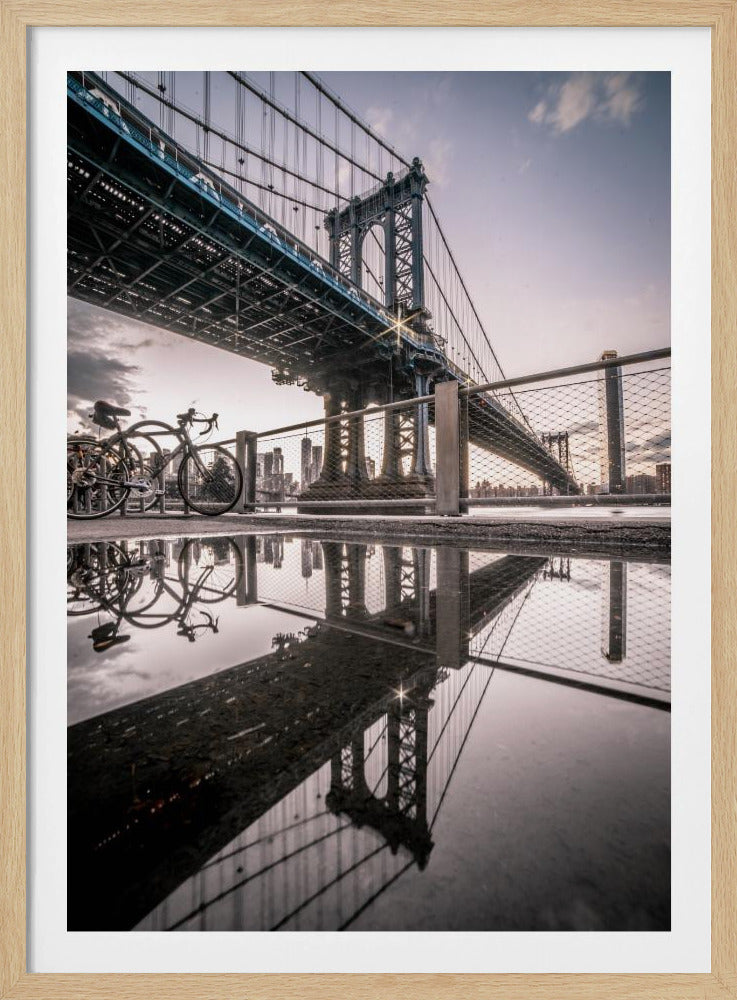 A dramatic low-angle photograph of the Manhattan Bridge, with its intricate steel structure and cables reflected in a large puddle on the pavement in the foreground. Two bicycles are parked near a fence on the left, with the New York City skyline visible in the distance under a softly lit, cloudy sky. Wall Art