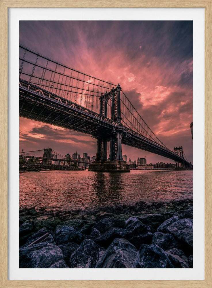 A dramatic low-angle view of the Manhattan Bridge over the East River at sunset. The sky is filled with pink and red clouds, which are reflected in the water below. The dark silhouette of the bridge contrasts with the vibrant sky, and the New York City skyline is visible in the background. In the foreground, dark, wet rocks line the riverbank. Decor