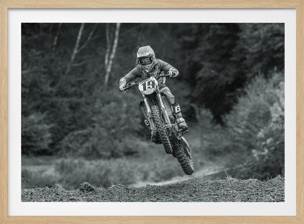 A dynamic black and white photograph of a motocross racer in mid-air, jumping over a dirt mound. The rider, wearing full protective gear and a helmet, is on a dirt bike with the number 19 plate. The background is a soft-focus forest, and the entire image is enclosed in a silver frame. Poster