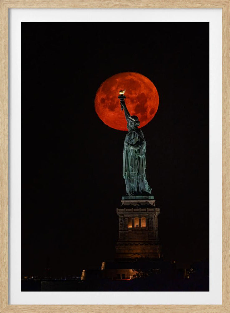 A striking night photograph of the Statue of Liberty perfectly silhouetted against a giant, vibrant red supermoon, which appears to be held aloft by the statue's torch against a deep black sky. Artwork